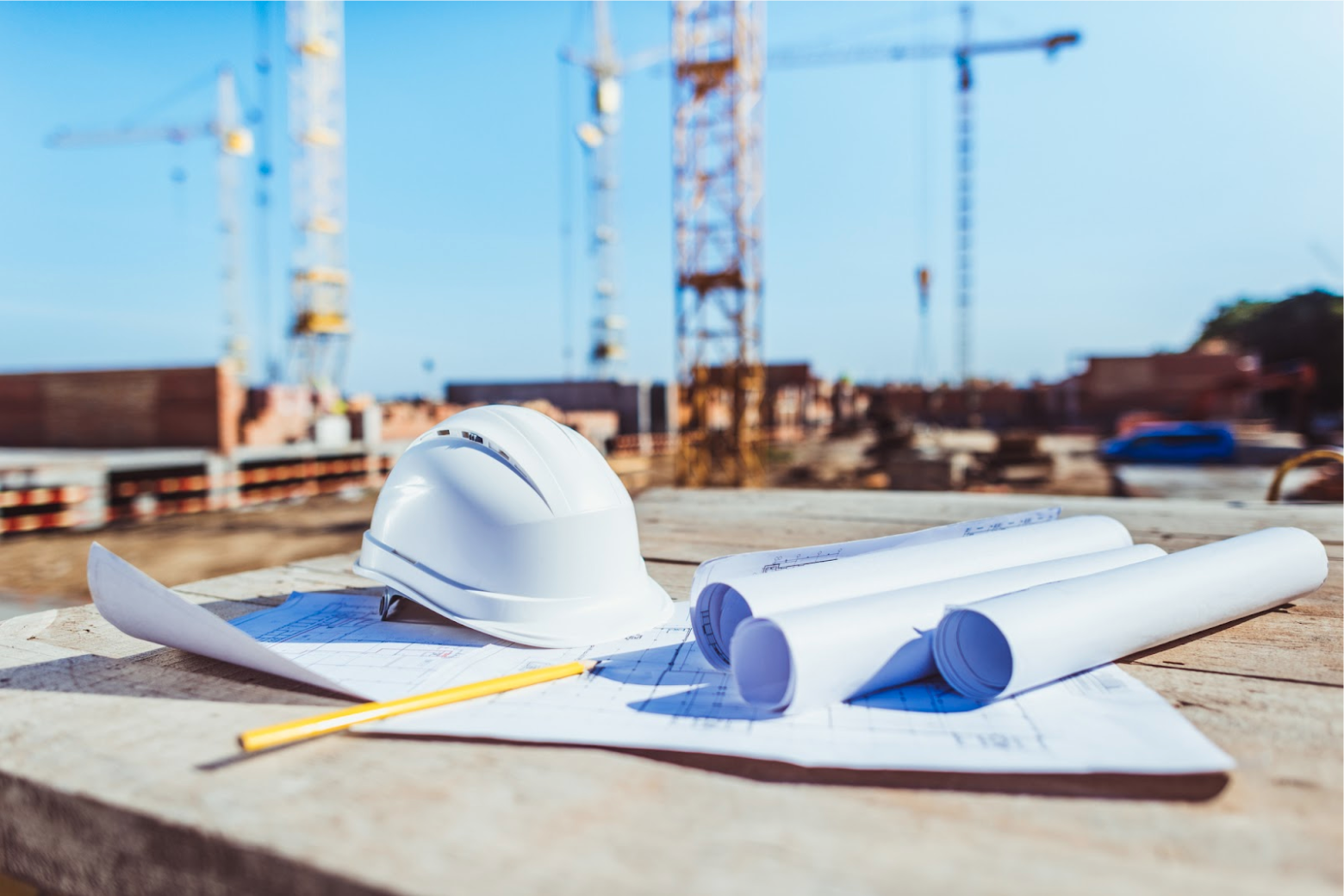 A white hardhat resting atop some blueprints, with three additional rolled-up blueprints to the side, and risers and cranes in a construction site visible in the background.